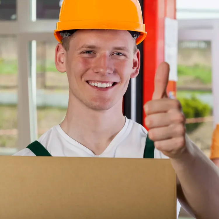 Ein junger Mann mit orangenen Helm haelt einen Karton in der einen Hand und haelt mit der anderen Hand den Daumen hoch