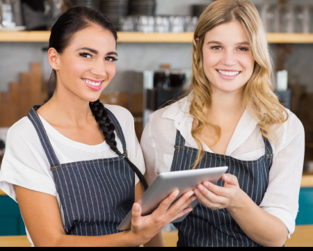 Zwei laechelnde Frauen in gestreiften Schuerzen, die in einem gastronomischen Umfeld arbeiten. Eine der Frauen haelt ein Tablet, waehrend sie gemeinsam in die Kamera schauen. Im Hintergrund sind Regale mit Geschirr und Kuechenutensilien zu sehen.