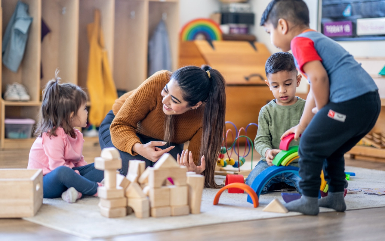 Auf dem Foto ist eine Erzieherin zu sehen, die mitten in Spielsachen kniet und mit Kindern um sich herum spielt. Sie hat braune Haare und ein braunes Oberteil an.